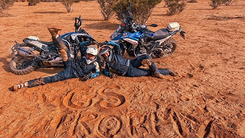 Uluslararası kadın takımından iki sürücü motosikletleri önünde kumda yatıyor, kumda “GS Trophy” yazısı yer alıyor.